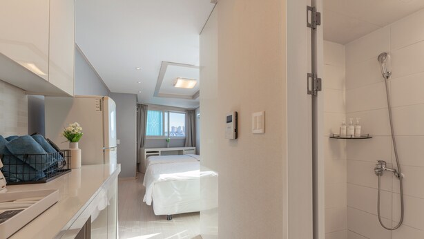 A modern hotel room with a bed, a bedside table with towels, a shower area, and a view of the city through the window. - Staywell Hotel (Busan)