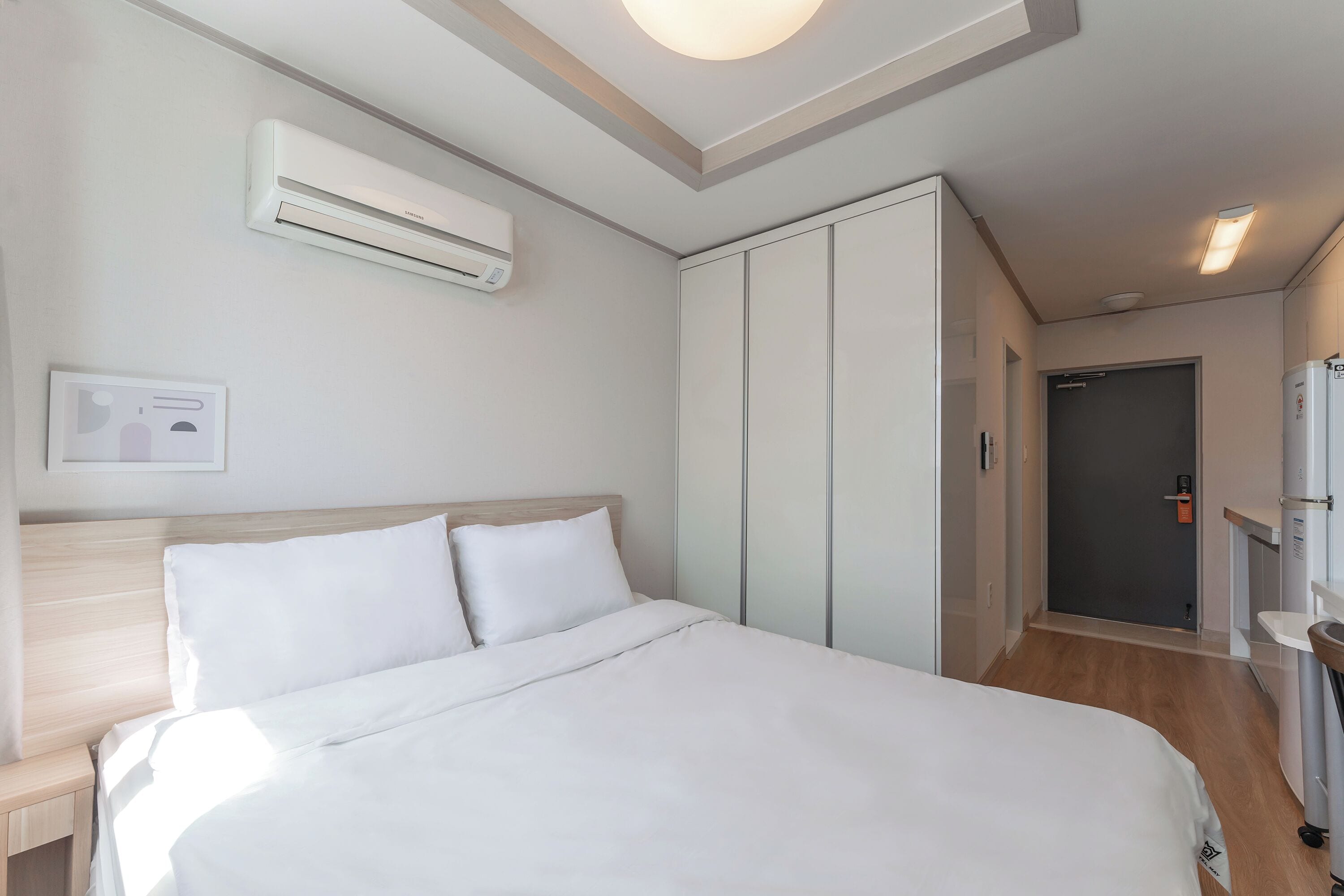 A hotel room with a bed, a wardrobe, an air conditioning unit, a bedside table, and a small bathroom area.
