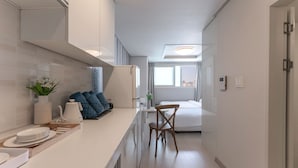 A modern hotel room with a kitchenette, a bed, a chair, and a view of the city. - Staywell Hotel (Busan)