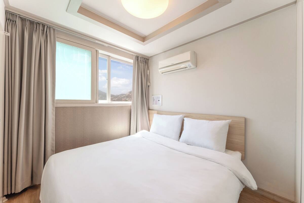 Habitación de hotel con una cama grande, una ventana con cortinas, aire acondicionado y vistas a la ciudad.