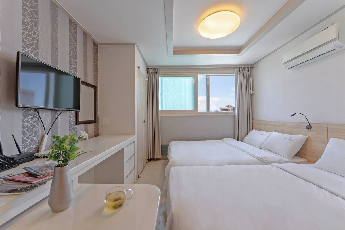 Habitación de hotel con televisión, escritorio con un florero y un vaso, dos camas, ventana con cortinas y un equipo de aire acondicionado.