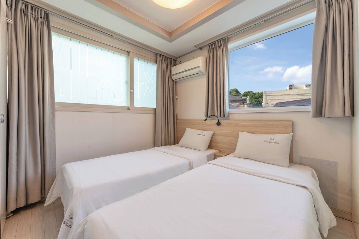 Habitación de hotel con dos camas, cortinas color beige, un equipo de aire acondicionado y vista a la ciudad.