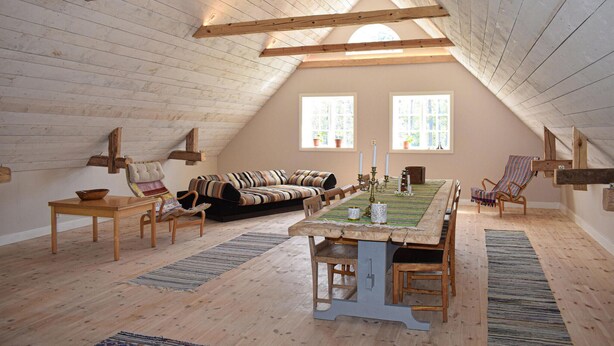 Une pièce mansardée chaleureuse avec un plafond haut, des poutres en bois, un canapé, une table à manger et deux fenêtres.