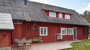 Une maison en bois rouge avec un toit en pente, une terrasse en bois avec des chaises et une fenêtre dotée d’un cadre blanc.