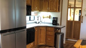 Una cocina compacta con armarios de madera, un refrigerador de acero inoxidable, una cafetera y un cubo de basura.