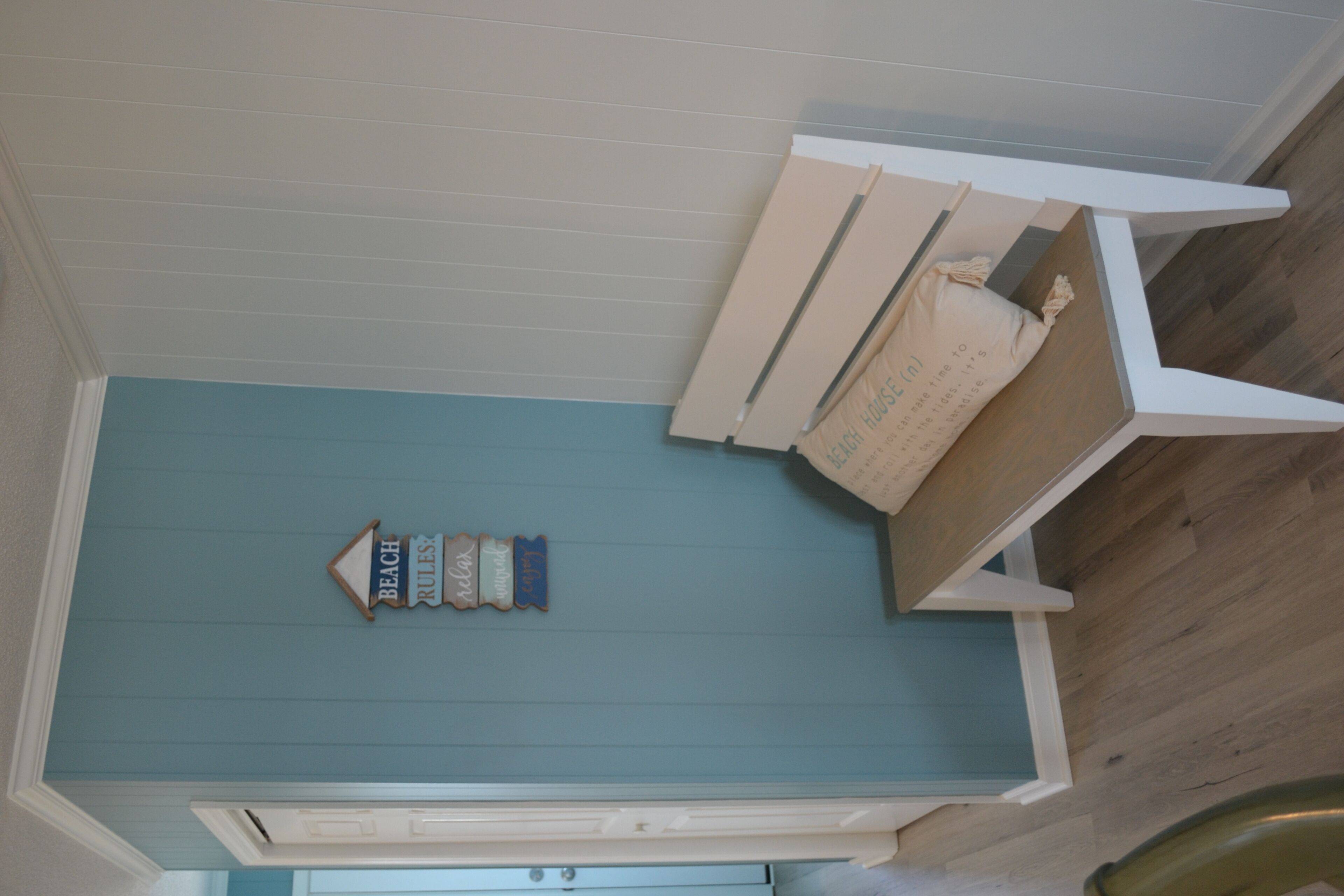 Una habitación con piso color turquesa, paredes blancas y detalles de madera. Se observa un mueble blanco con espejo y una pequeña repisa decorada.