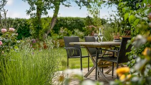 An outdoor dining area with a wooden table and chairs, surrounded by lush greenery and flowering plants. - Gorgeous home in Sydals with WiFi (Sydals)