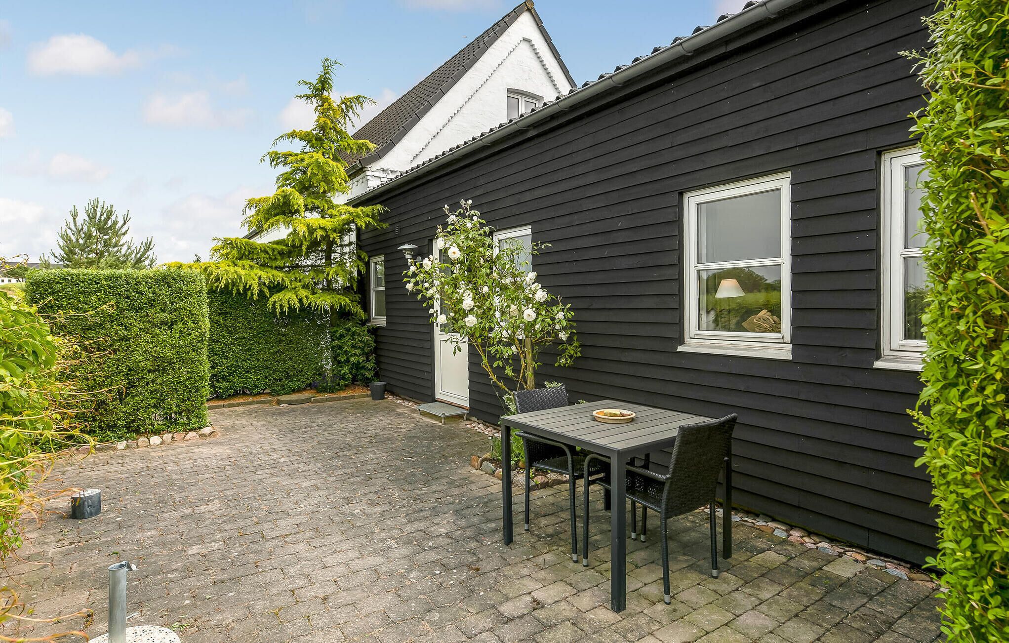Une maison noire avec un patio dallé, une table entourée de chaises et un petit jardin.