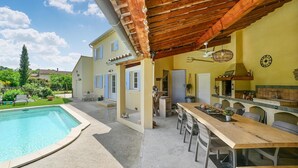 Zona junto a la piscina con una pérgola de madera, mobiliario exterior y una cocina con un reloj y un letrero que dice "GARDEN".