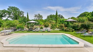 Una piscina rectangular con borde de azulejos, rodeada por una solana de hormigón. Hay sillas de descanso y sombrillas dispuestas alrededor de la piscina, y una valla de madera al fondo.