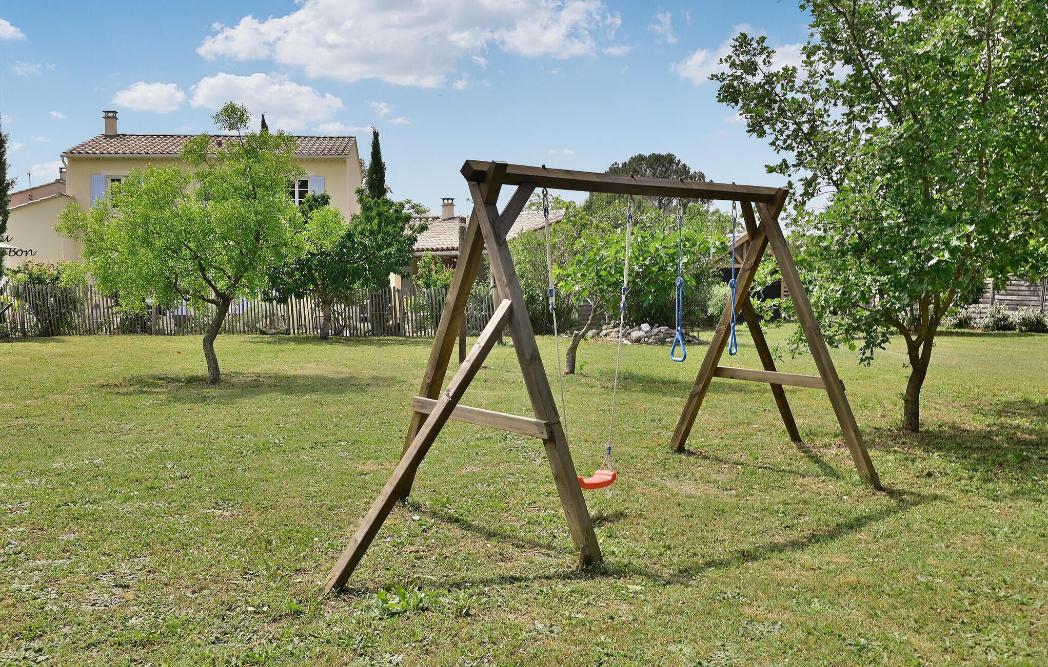 A wooden swing set in a grassy yard with trees and a house in the background.