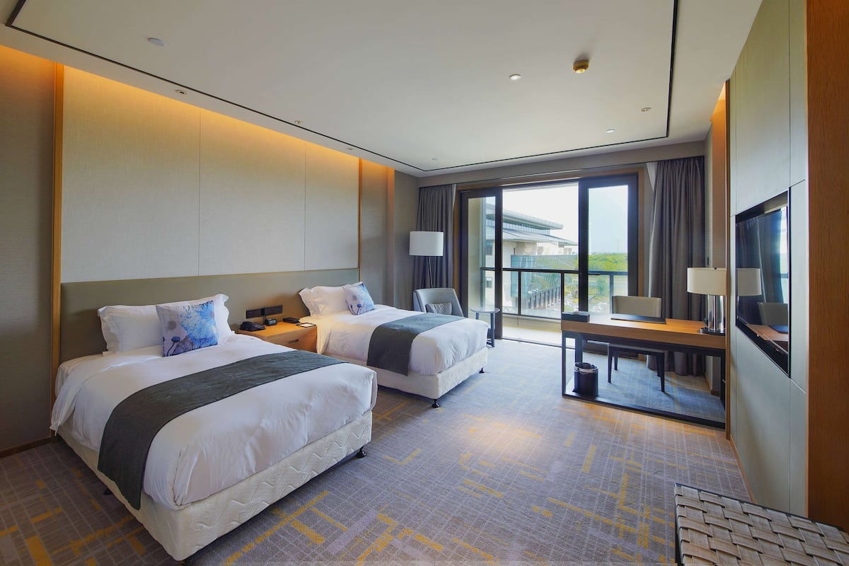 A hotel room with two beds, a desk, a chair, and a large window offering a view.