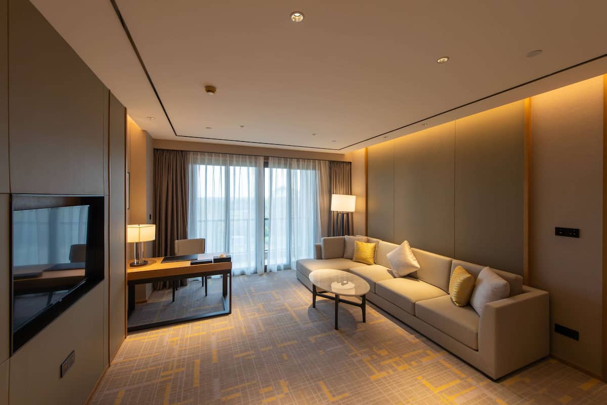 A modern hotel room with a sofa, a coffee table, a TV, and a large window with curtains.