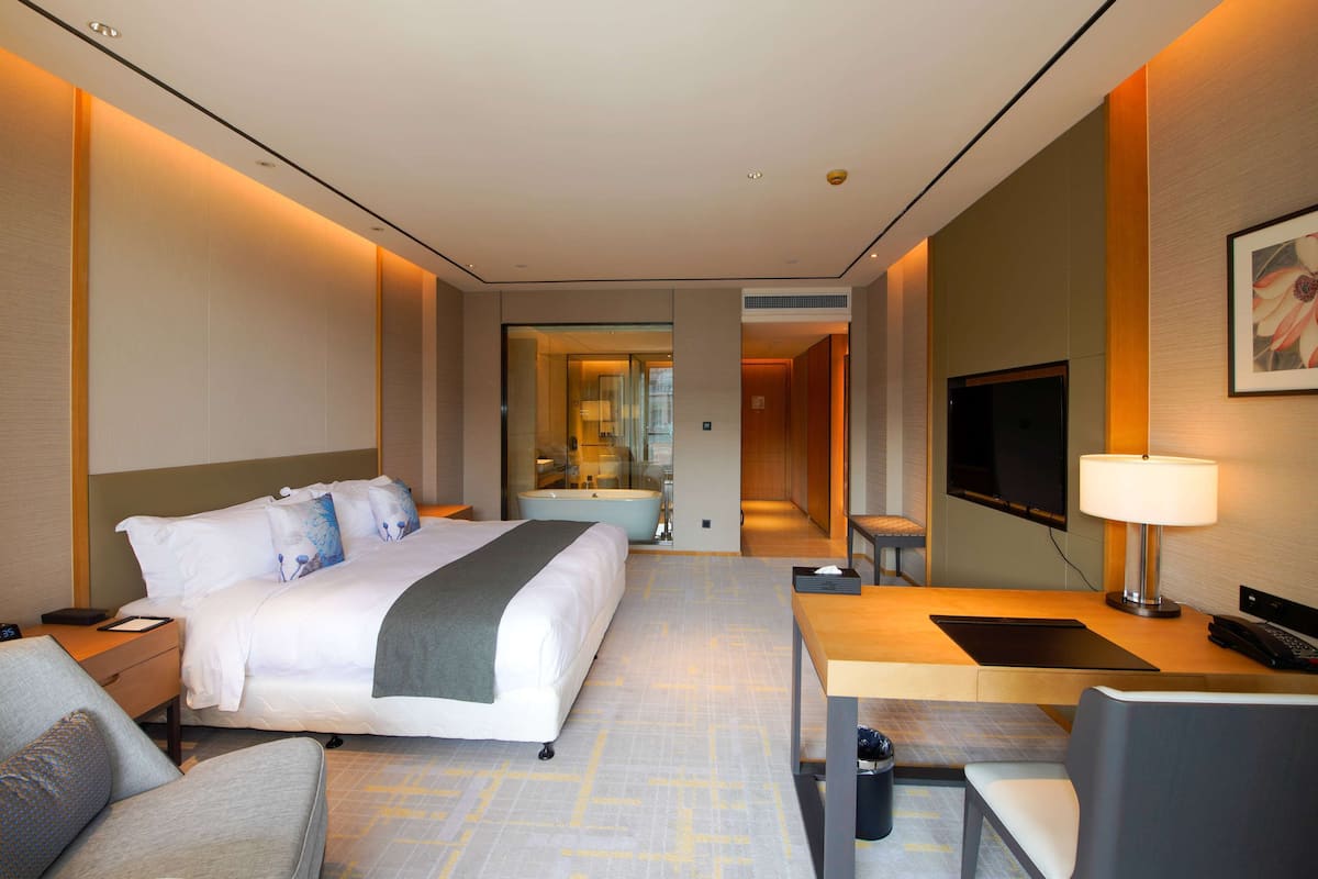 A hotel room with a bed, bedside table, lamp, TV, and a desk with a chair.