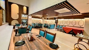 Um restaurante moderno com mesas de madeira e cadeiras azuis, teto alto com ripas de madeira, e um bar ao fundo.