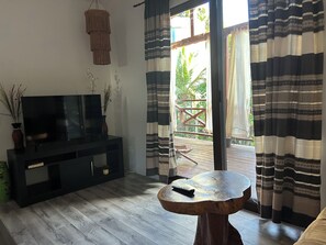 A room with a flat-screen TV on a black stand, a wooden coffee table with a remote, and a sliding glass door leading to an outdoor area.