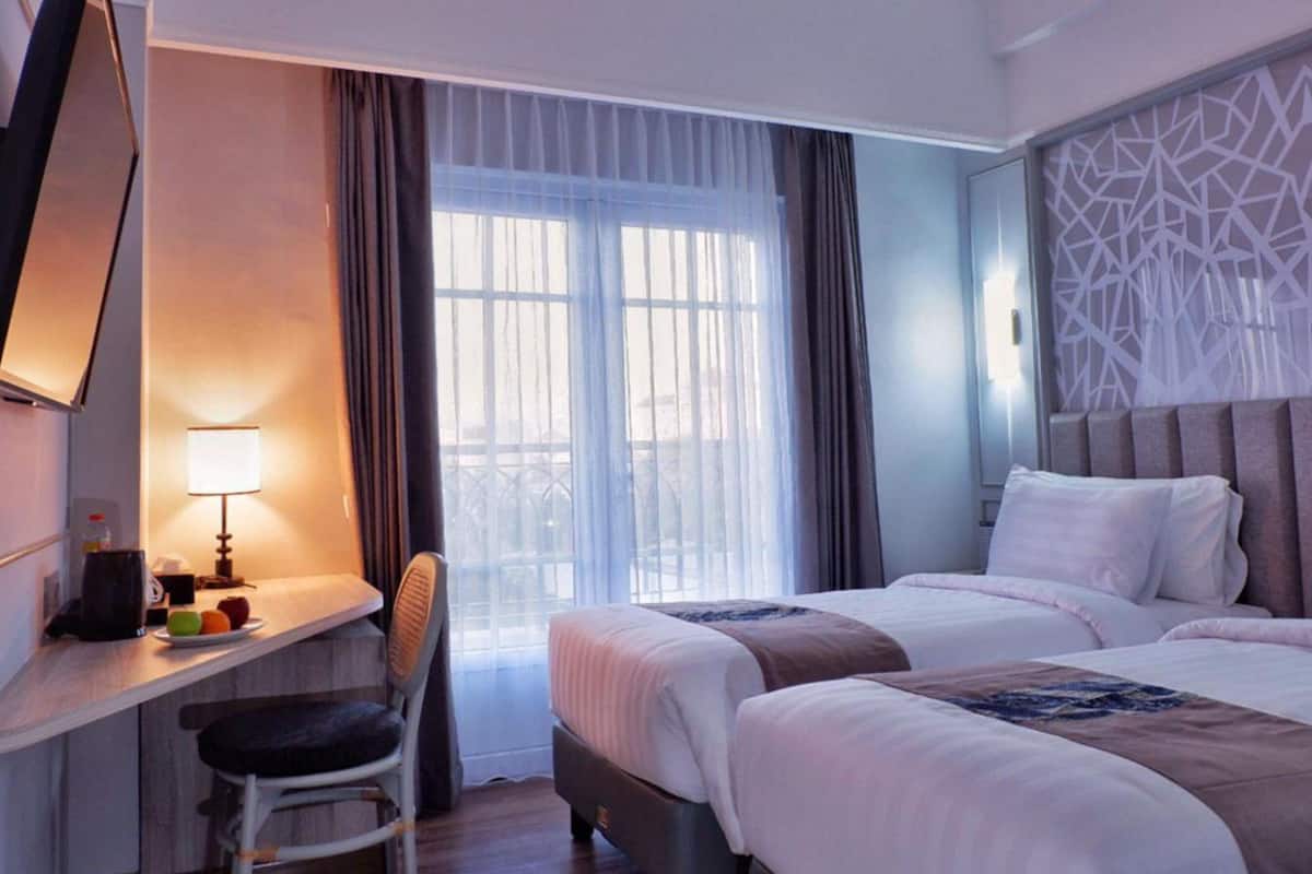 Hotelkamer met twee bedden, een bureau met stoel, een raam met dunne gordijnen en een decoratief wandpaneel.