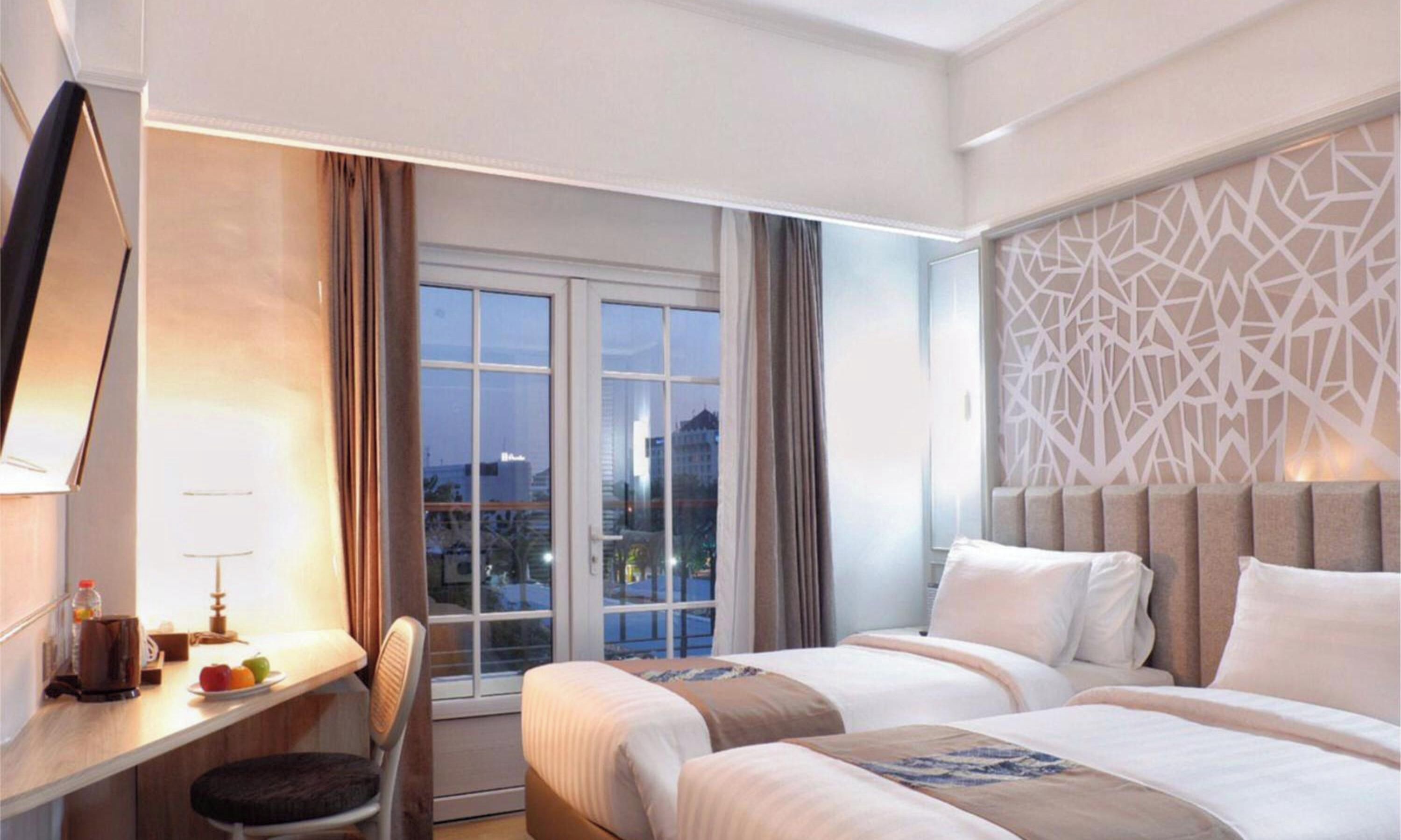 A hotel room with a desk, two beds, a large window, and a decorative wall panel.
