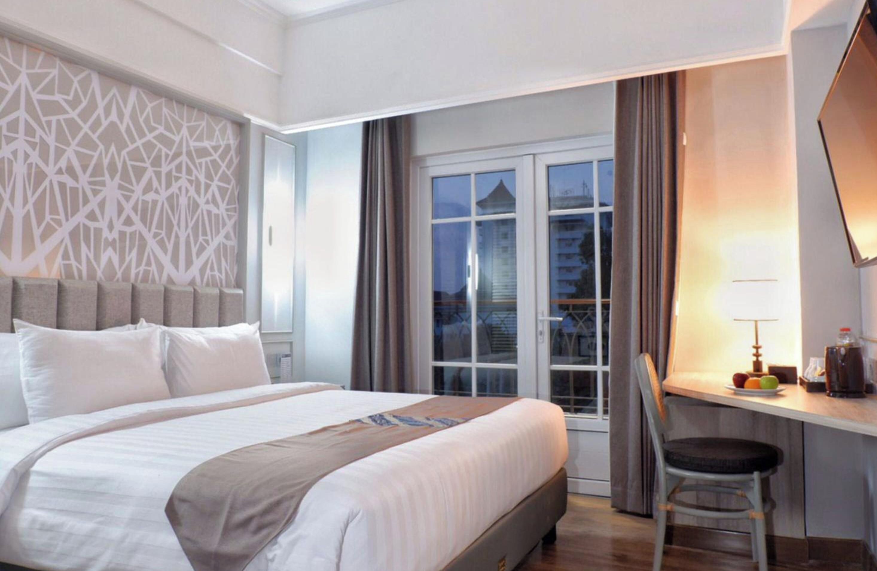 A hotel room with a bed, a desk, a chair, and a window with a view of buildings.