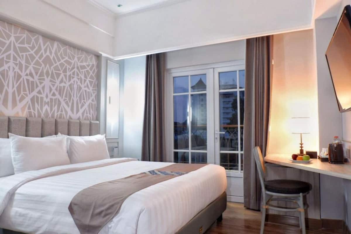 Hotelkamer met een groot bed, een bureau met stoel, een raam met gordijnen en uitzicht op gebouwen.