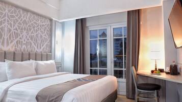 A hotel room with a large bed, a desk with a chair, a window with curtains, and a view of buildings outside.