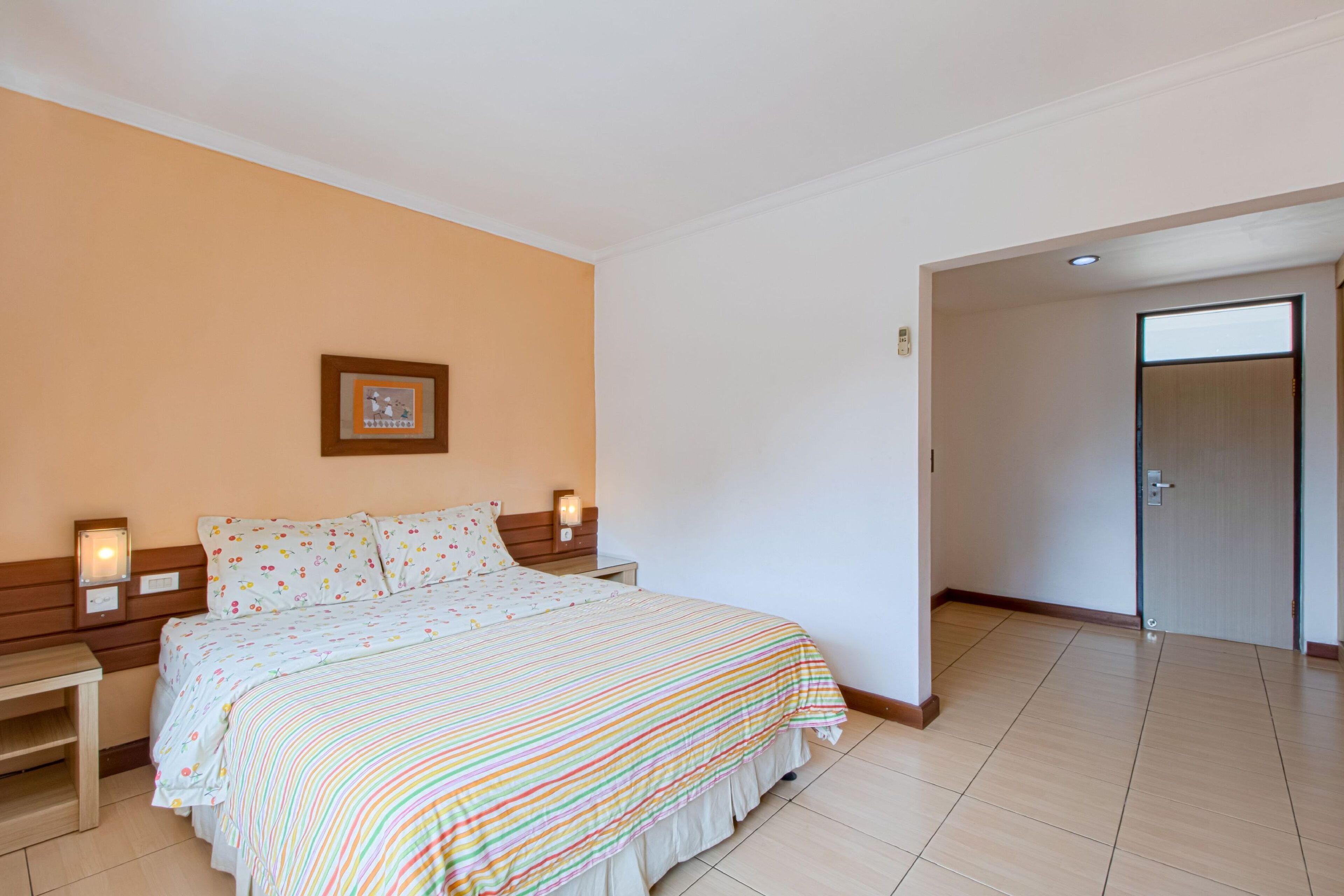 A hotel room with a bed, bedside tables, a framed picture on the wall, and a door leading to another room.