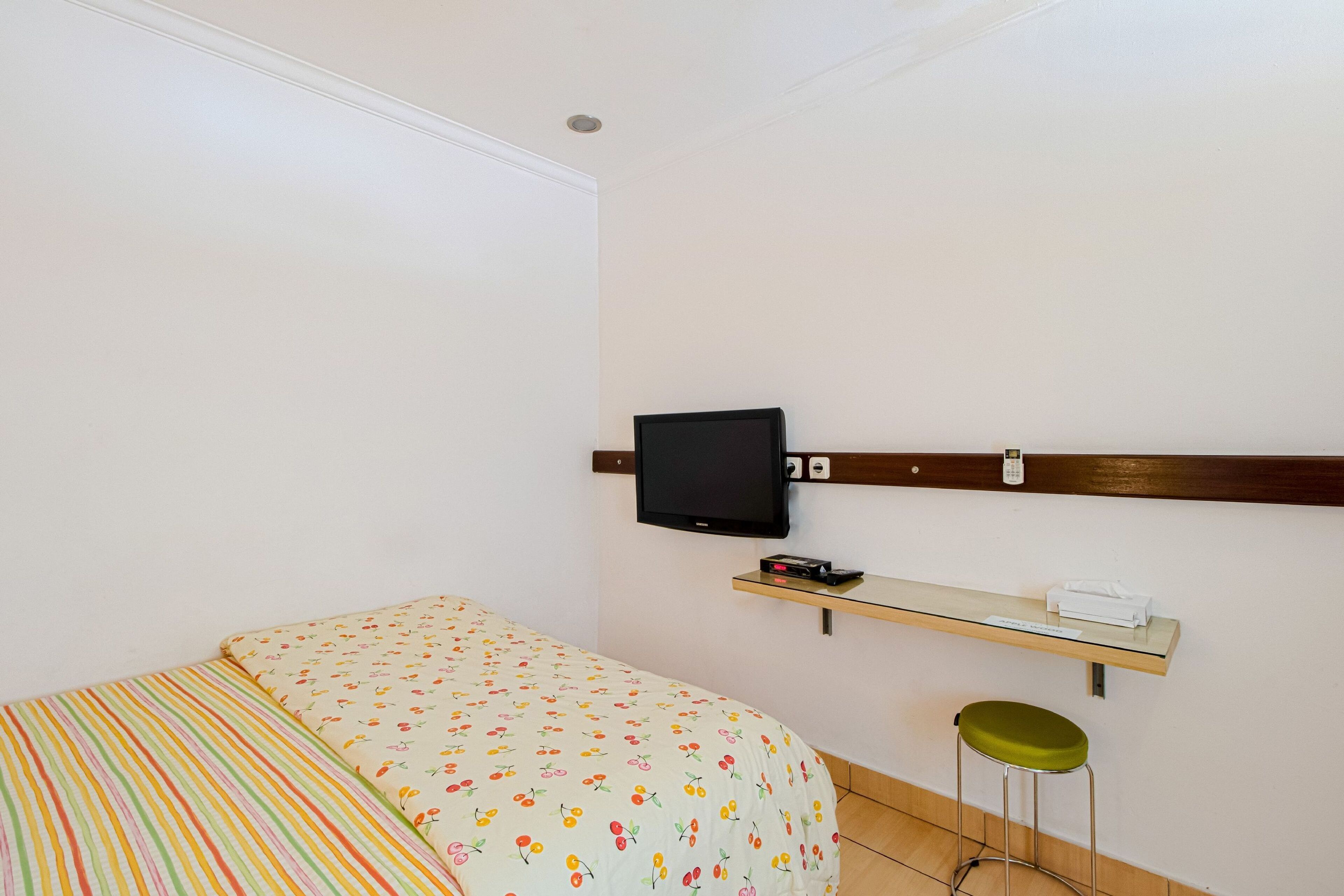 Quarto com cama, televisão na parede, prateleira pequena com telefone e um banquinho.