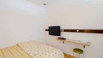 Quarto com cama, televisão na parede, prateleira pequena com telefone e um banquinho.