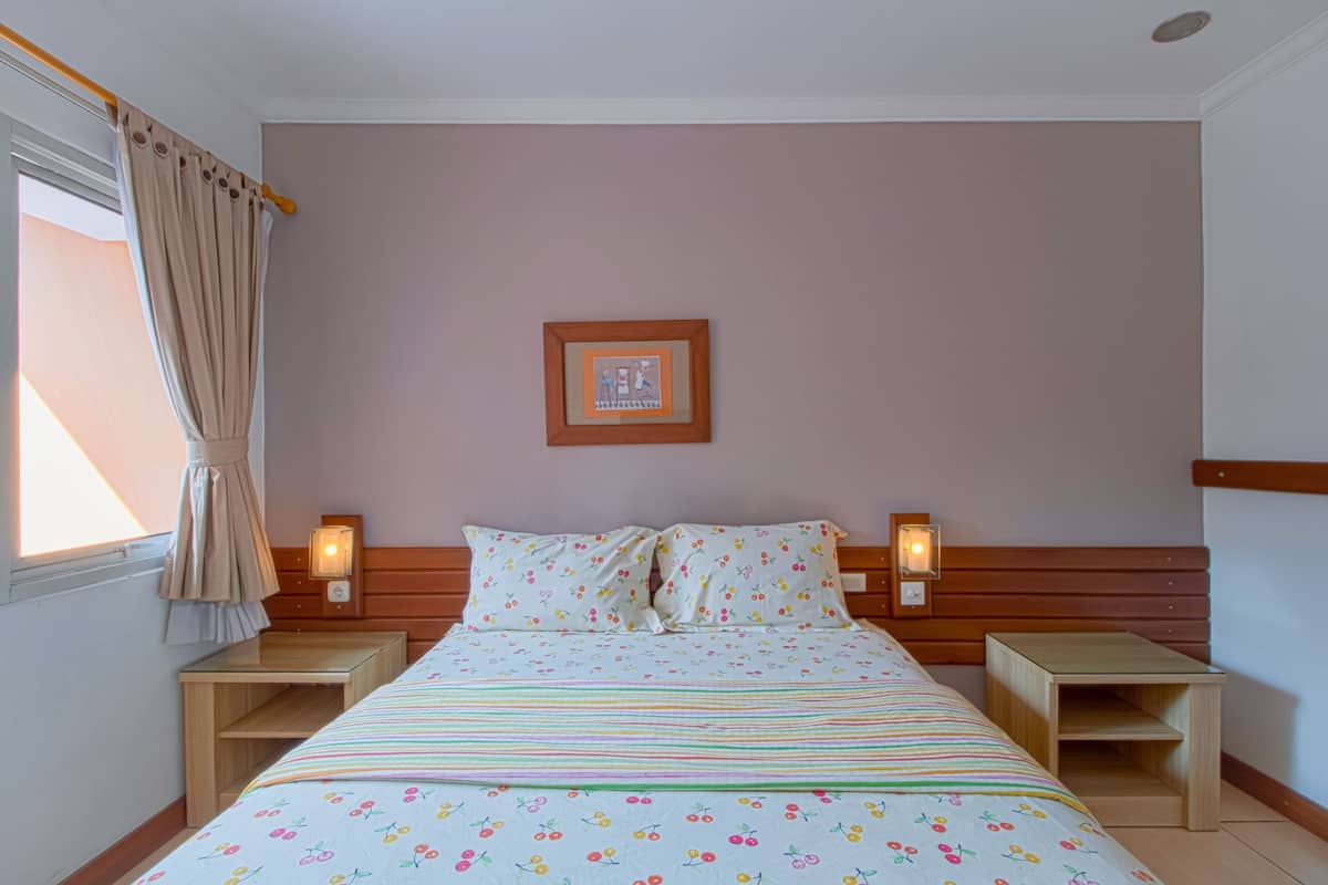 Un letto rifatto con cura, una coperta a motivi floreali, due comodini con lampade, un quadro appeso al muro e una finestra con tende.