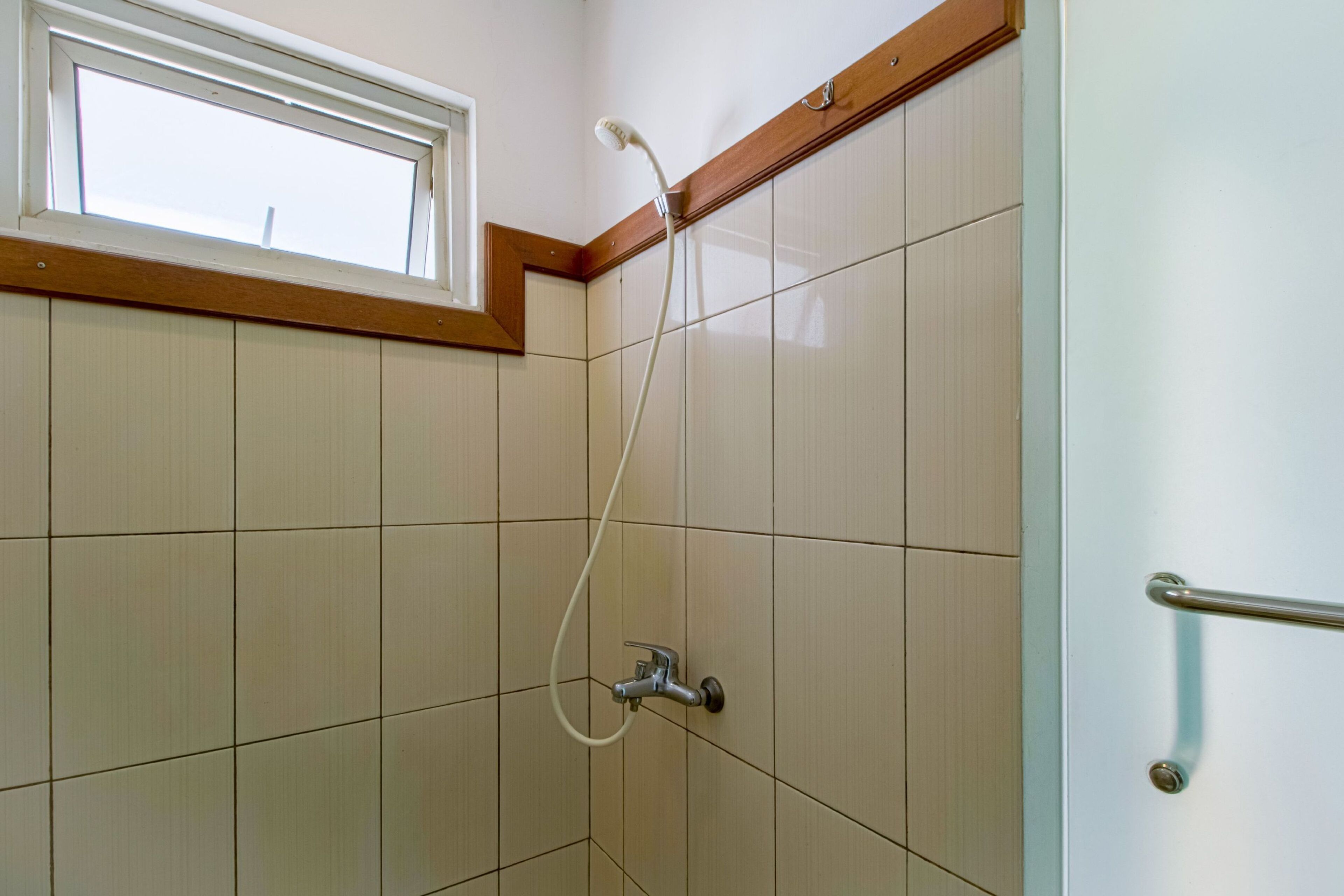 A bathroom with beige tiled walls, a window, and a shower with a handheld showerhead.