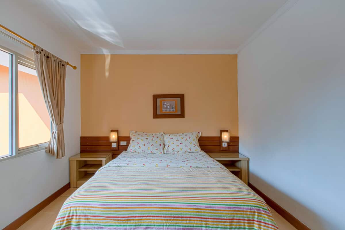 Una camera d'albergo con un letto, due comodini, un tavolo di fianco al letto, una finestra con tende e un quadro appeso al muro.