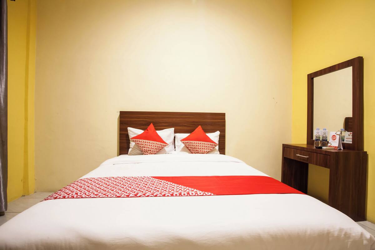 A hotel room with a bed, two red pillows, a wooden headboard, a nightstand, and a mirror.