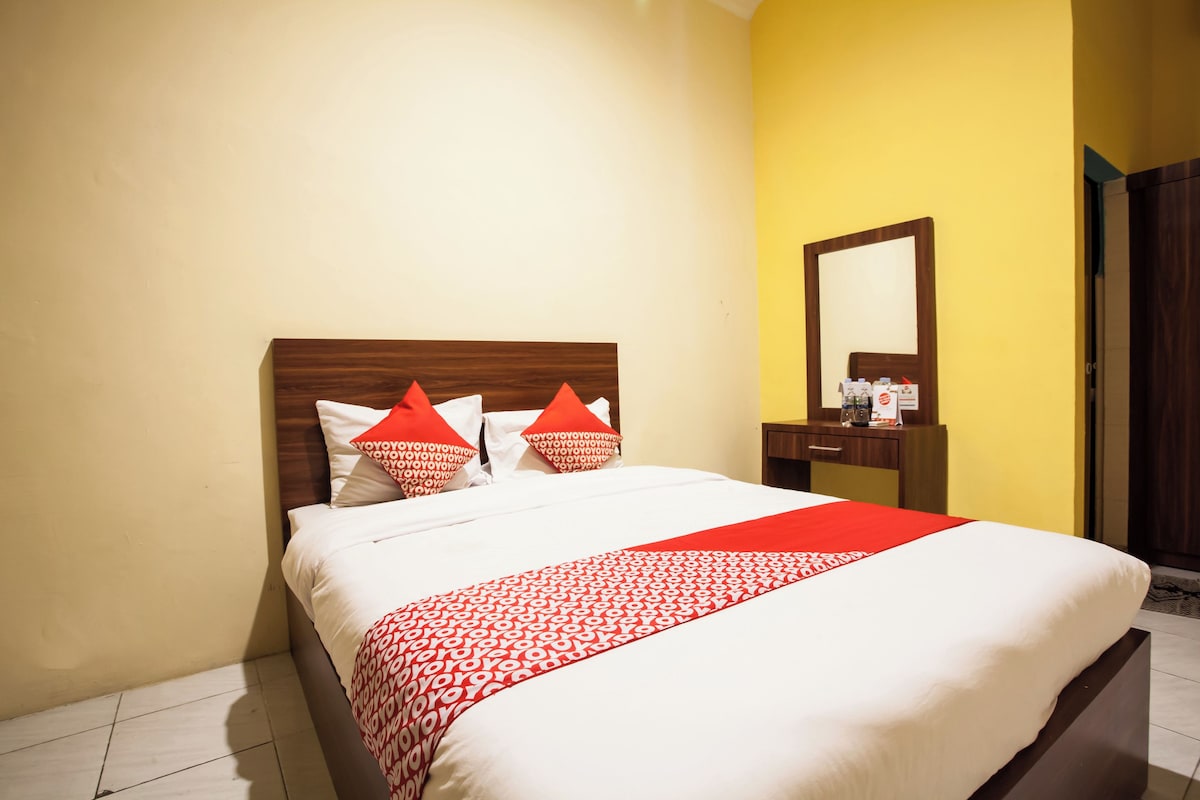 A neatly made bed with a red and white patterned bedspread and pillows, a wooden headboard, and a small bedside table with a mirror and some items on it.