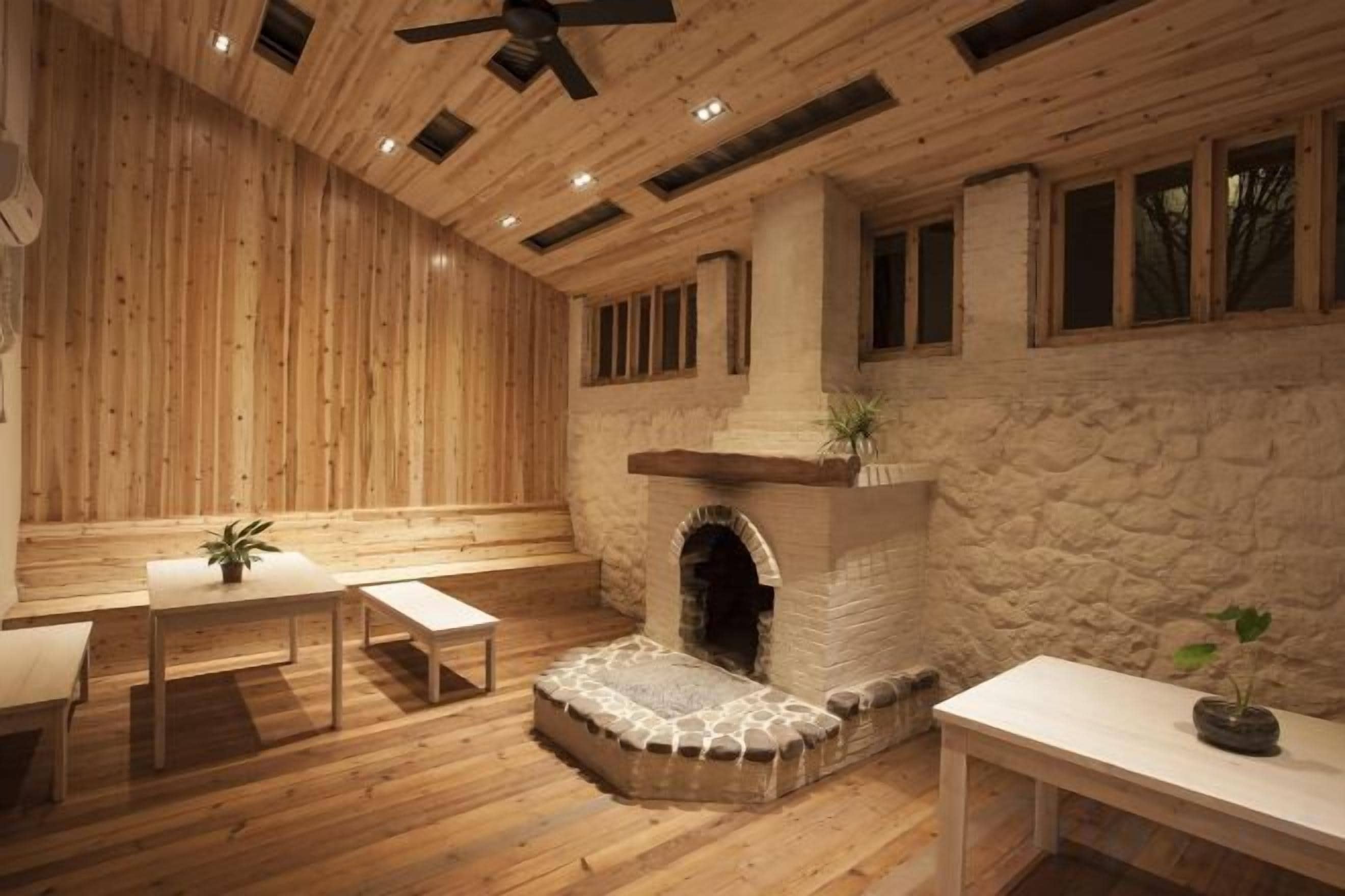 A room with wooden walls, a stone fireplace, a ceiling fan, and a table with a bench.