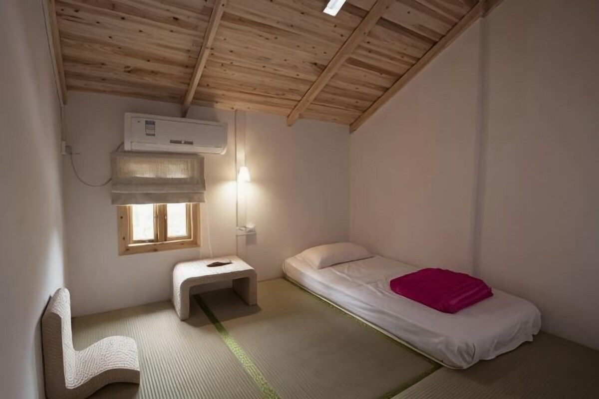 A small, single-room accommodation with a wooden ceiling, a bed, a chair, a small table, and a window.