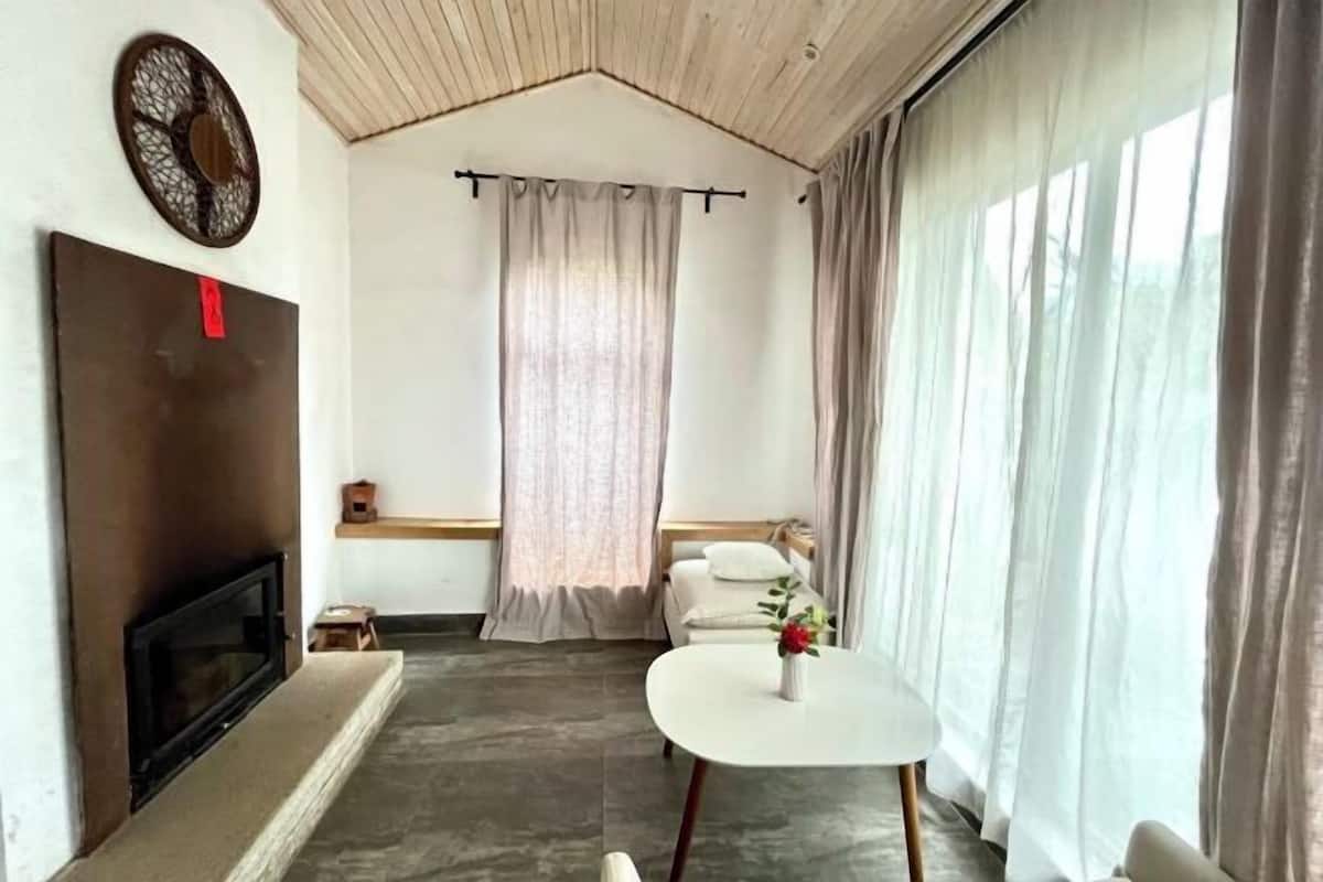 A room with a fireplace, a small table with a vase, a bed with pillows, and a large window with curtains.