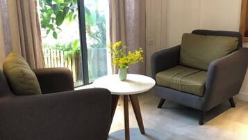Una sala de estar moderna con un sofá y un sillón, una mesita redonda con un jarrón de flores, y un ventanal con cortinas.