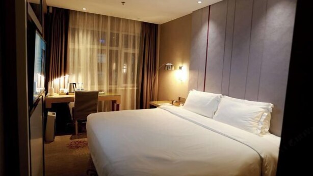 A hotel room with a large bed, a desk, and a chair. There is a window with curtains and a wall-mounted lamp. - Lavande Hotel (Zhuanghe Xiangyang Road) (Zhuanghe)