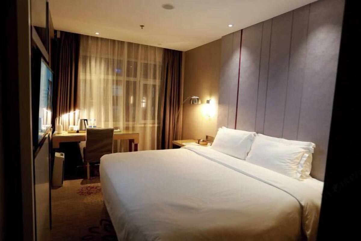 A hotel room with a large bed, a desk, and a chair. There is a window with curtains and a wall-mounted lamp.
