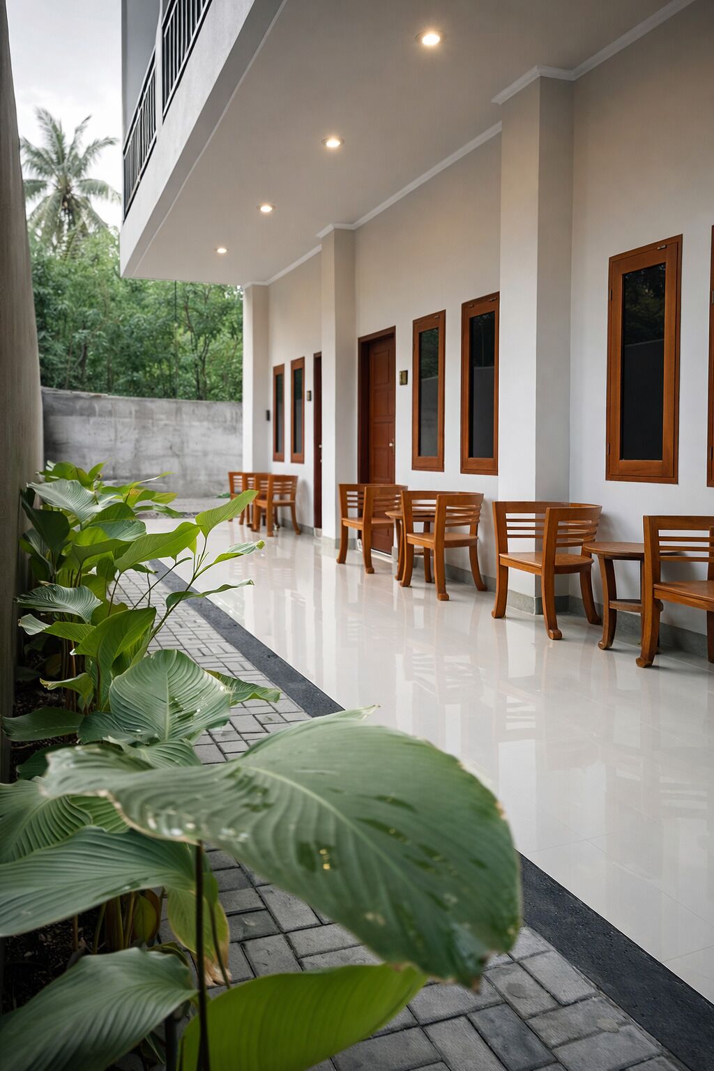 A covered outdoor seating area with wooden chairs and tables, surrounded by large windows and a concrete wall.