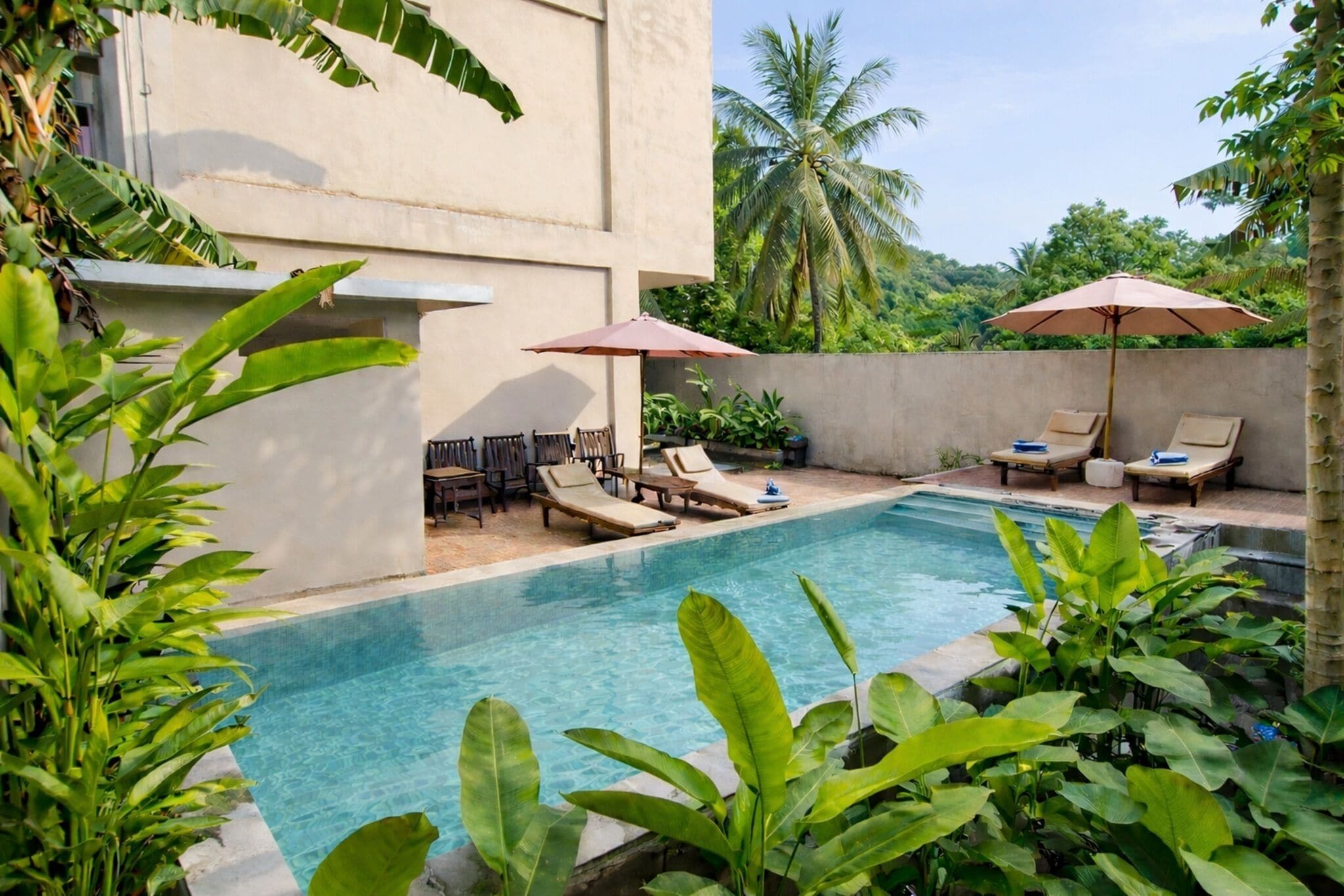 A pool area with lounge chairs and umbrellas, surrounded by lush greenery and palm trees.