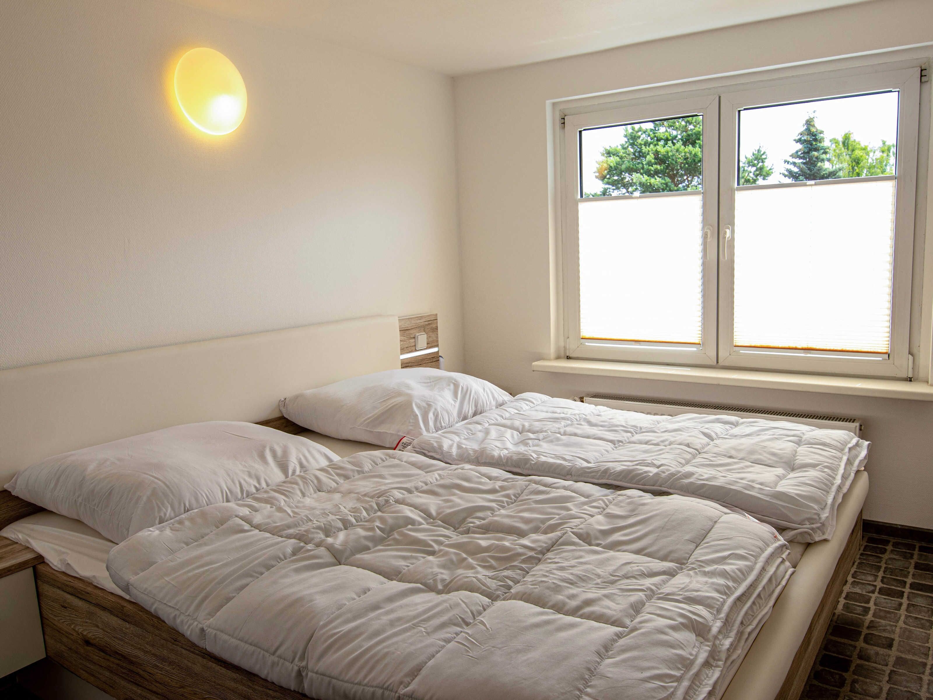 Un dormitorio con cama, mesita de noche, lámpara de pared y ventana con persianas.