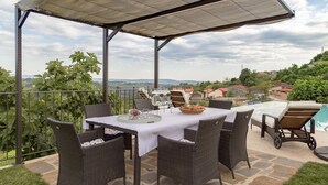 En uteplats med möbler i rotting, ett bord dukat för måltider och utsikt över en swimmingpool och det omgivande landskapet.