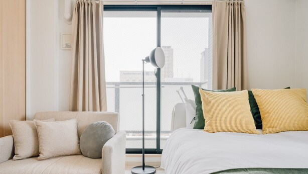 Habitación de hotel con cama, sofá, lámpara de pie y vista a un paisaje urbano a través de un ventanal grande.