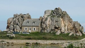 Una casa de piedra con techo inclinado, ubicada en una colina rocosa con una masa de agua en primer plano.