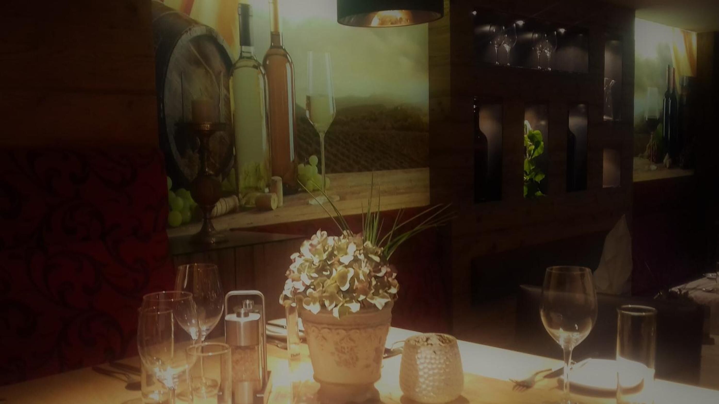 Una mesa con copas de vino, una botella y una planta en maceta en una habitación con poca luz y decoración de madera.