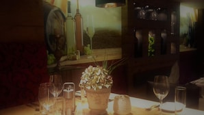 A table with wine glasses, a bottle, and a potted plant in a dimly lit room with wooden decor. - Das kleine Hotel (Stumm)