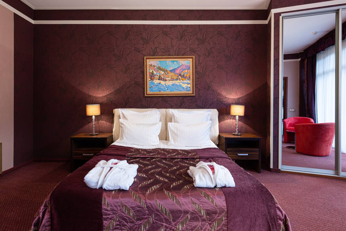 A hotel room with a large bed, two bedside tables with lamps, a painting on the wall, and a red chair in the corner.