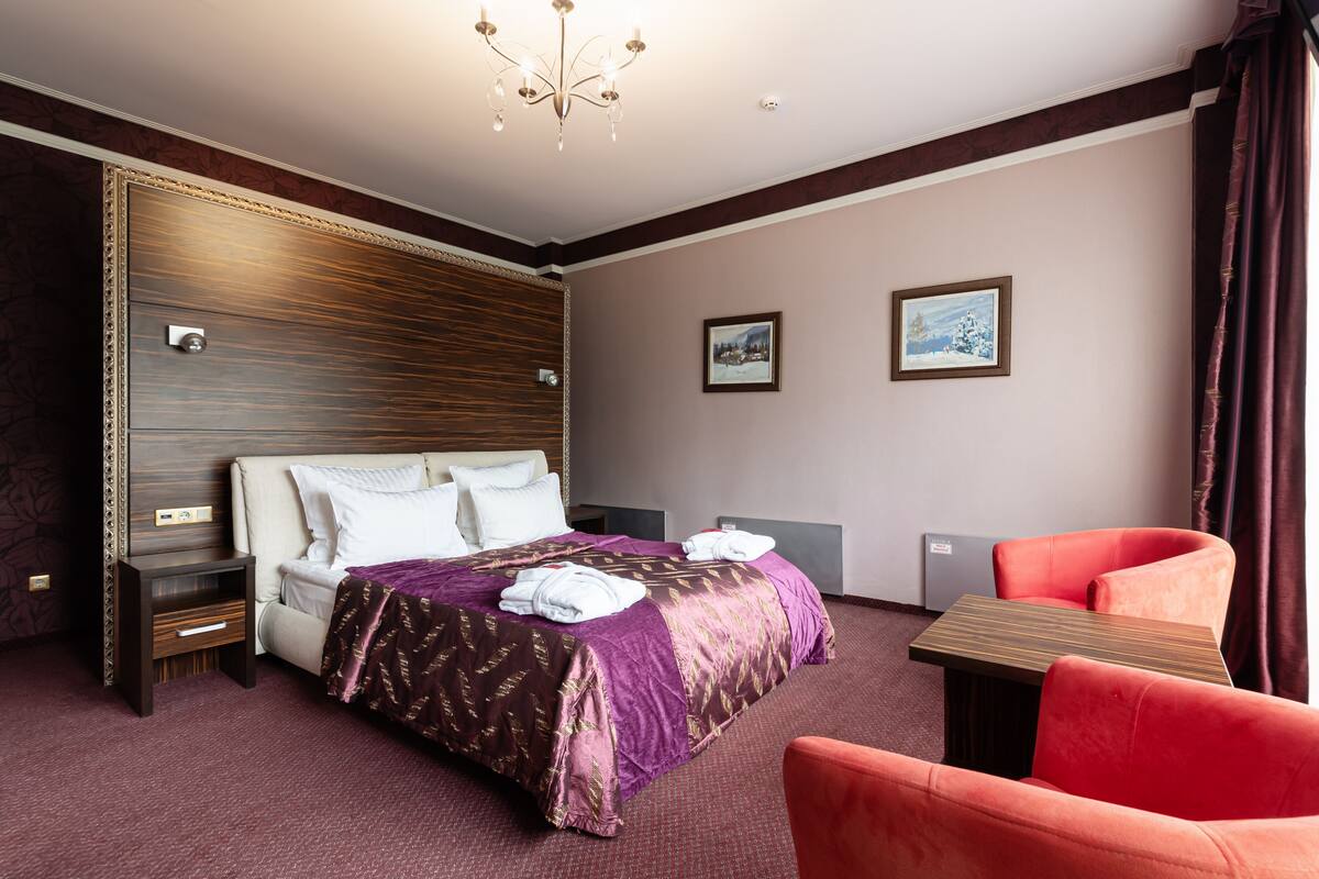 A hotel room with a large bed, a nightstand, a table, and a red chair.