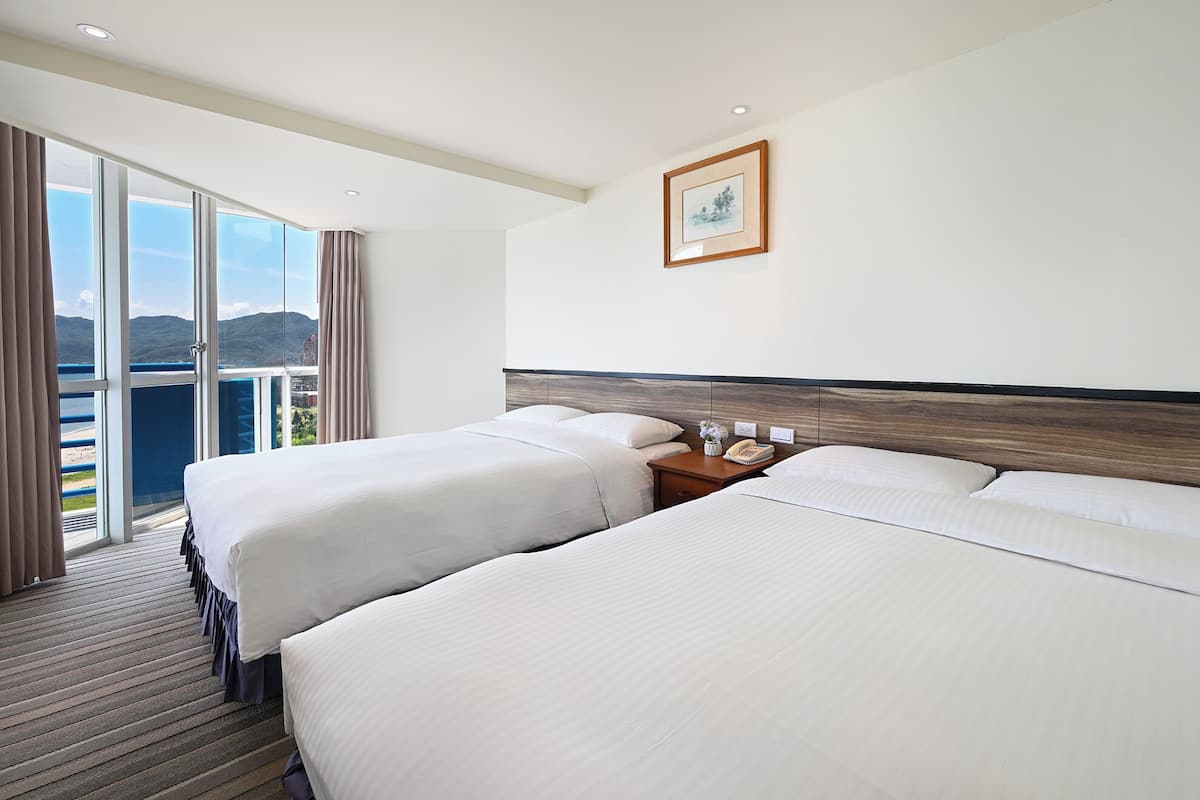 A hotel room with two beds, a wooden headboard, a nightstand with a phone, and a large window with a view of the mountains.